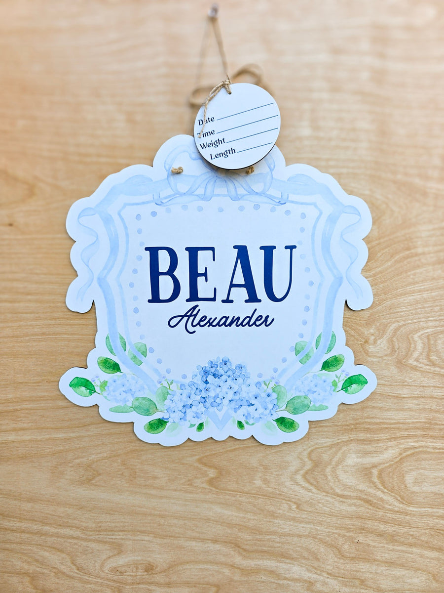 Personalized blue hydrangea and bow baby hospital door sign with navy lettering, green leaves and scalloped crest shape