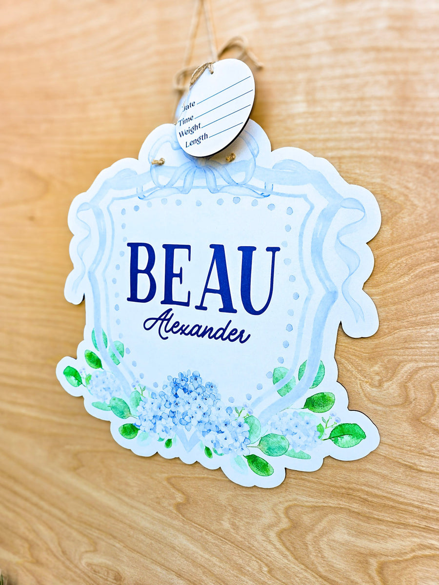 Angled view of personalized blue hydrangea hospital door hanger with watercolor bow, decorative crest and detachable birth stat round